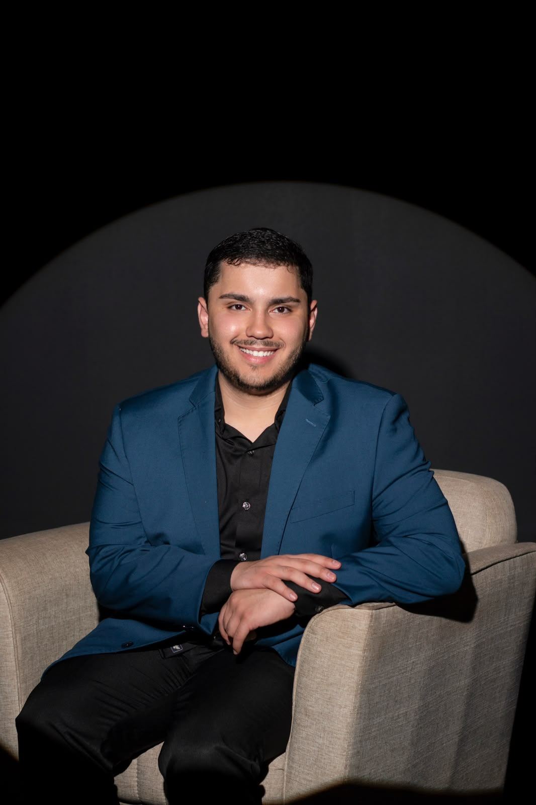 Marcus Trivellato sorrindo, sentado em uma poltrona clara, de blazer azul-marinho